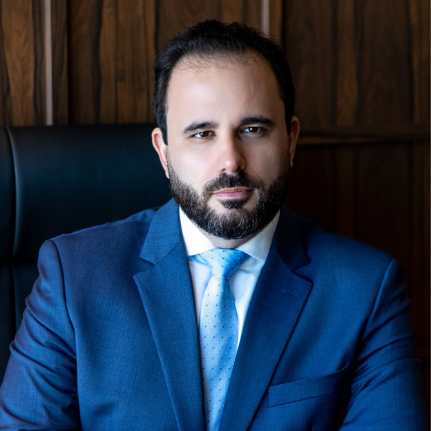 Retrato profissional de Guilherme Sombrio, sócio fundador do CR. Andrade & G.J. Sombrio, vestindo terno azul e gravata azul clara, com expressão séria e ambiente institucional ao fundo.
